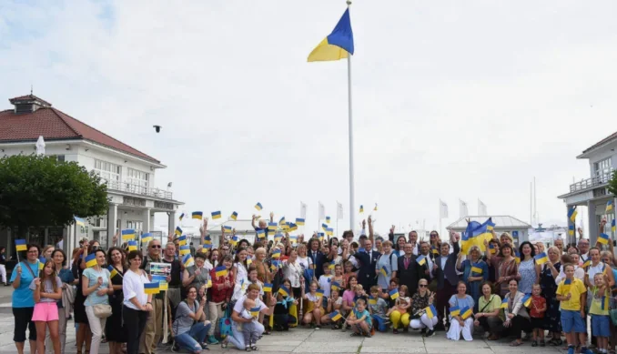 Толпа людей с многочисленными украинскими флагами возле белых зданий под большим флагом