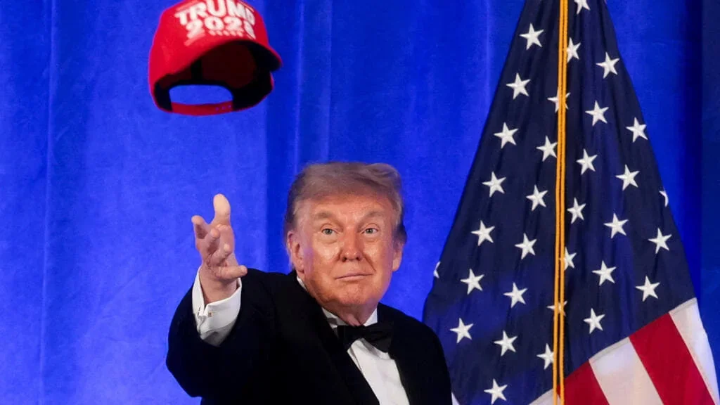 Дональд Трамп у смокінгу кидає червону кепку TRUMP 2028 на тлі прапора США