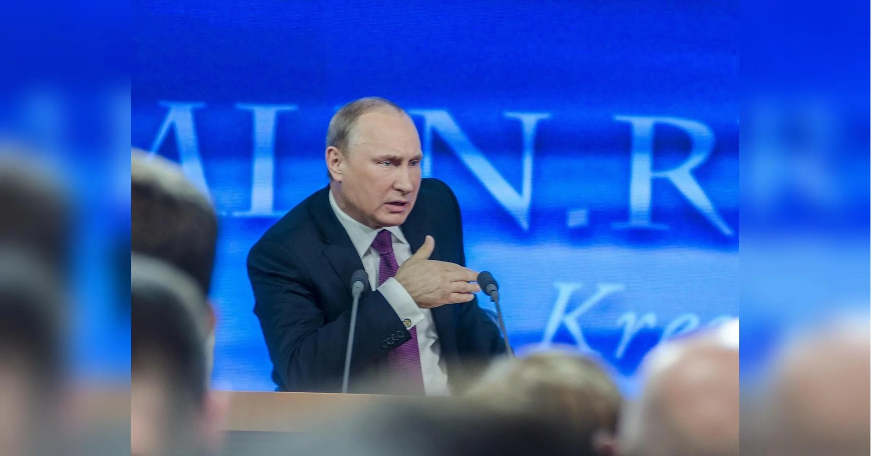 Путін жестикулює біля мікрофонів за трибуною на тлі синього екрана