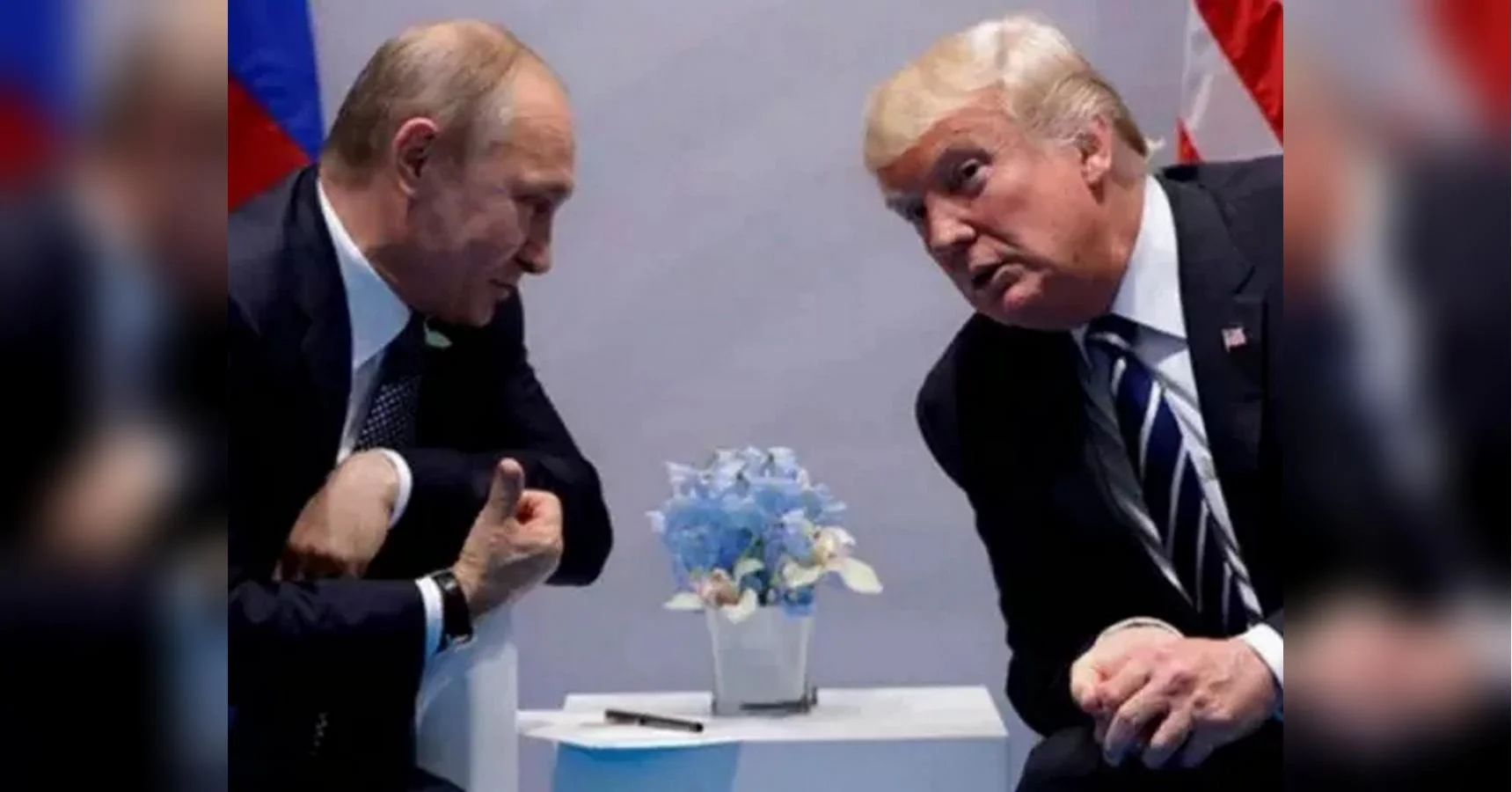 Путін і Трамп сидять за столом із блакитними квітами на тлі прапорів