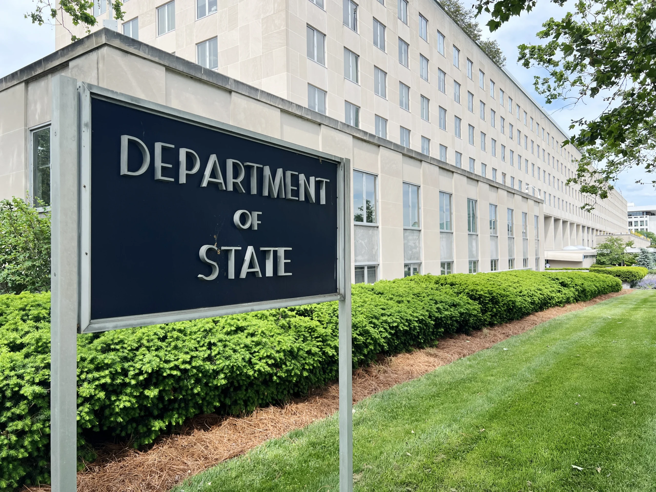 Вивіска Department of State перед будівлею Держдепу США серед зелених кущів