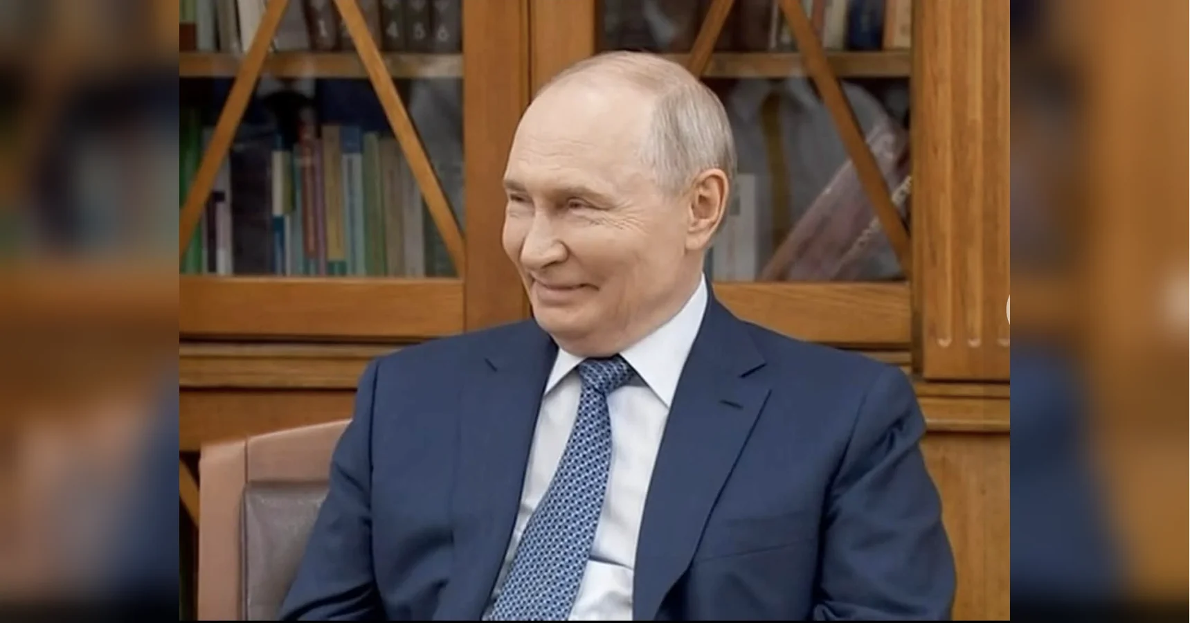Володимир Путін у синьому костюмі сидить і посміхається перед книжковою шафою