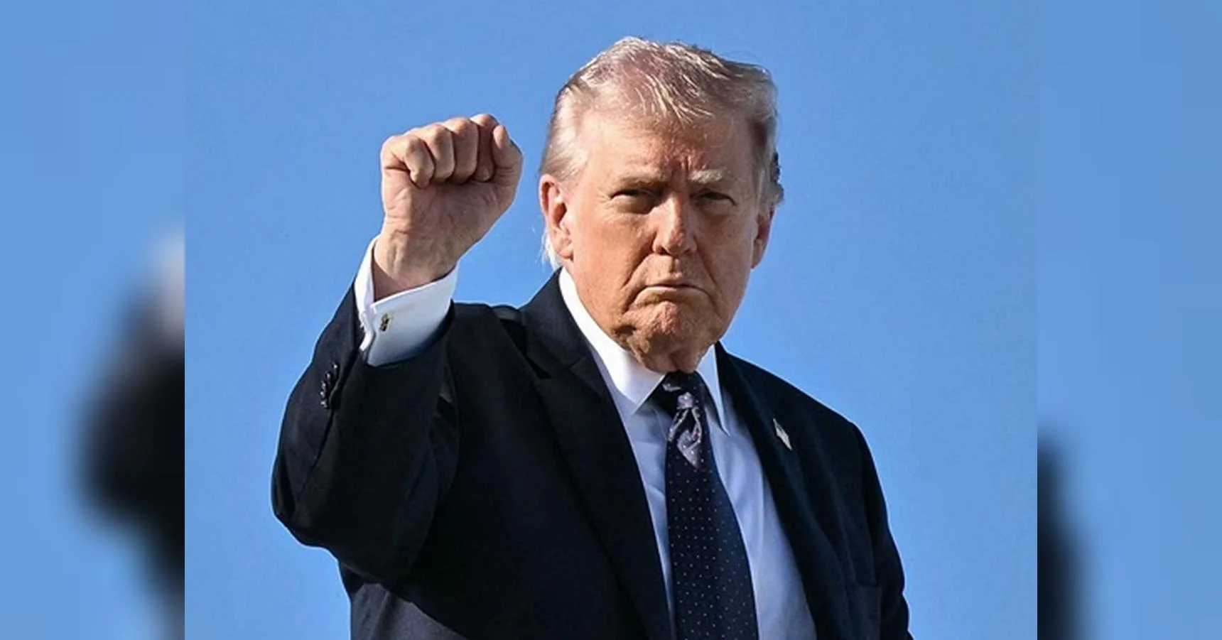 Дональд Трамп у костюмі піднімає кулак на тлі блакитного неба