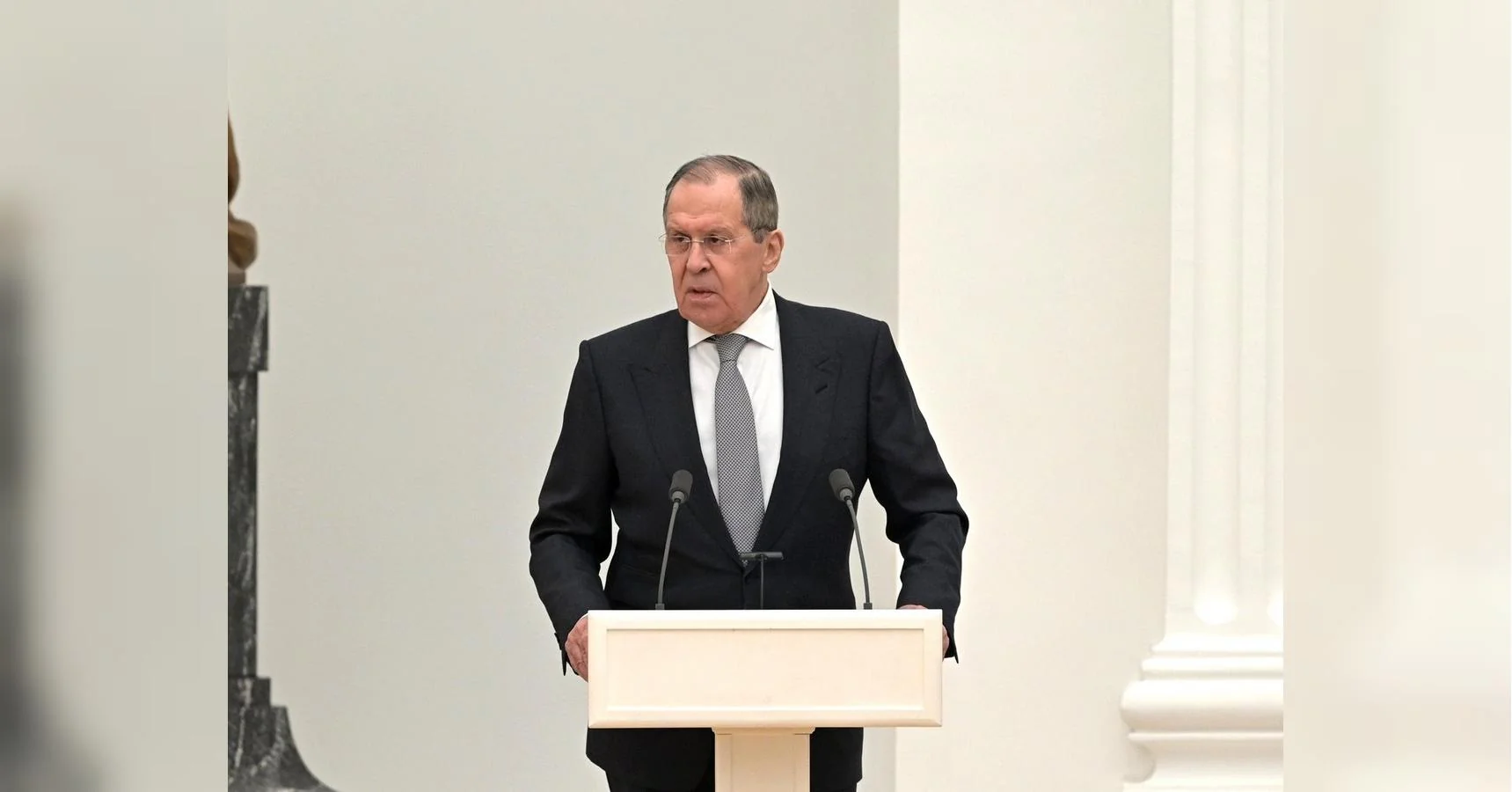 Сергій Лавров у костюмі виступає за білою трибуною з мікрофонами
