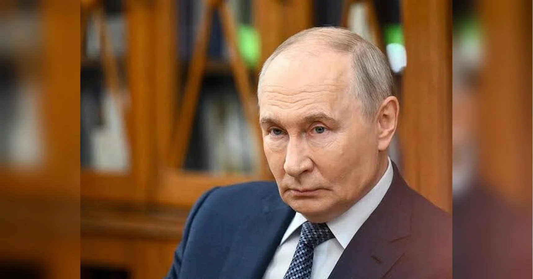 Володимир Путін у темному костюмі серйозно дивиться вбік на тлі дерев’яних книжкових шаф